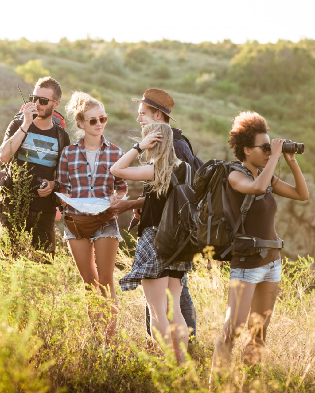 Four friends hiking through tall grass in a sunny field, two using binoculars and one holding a map.