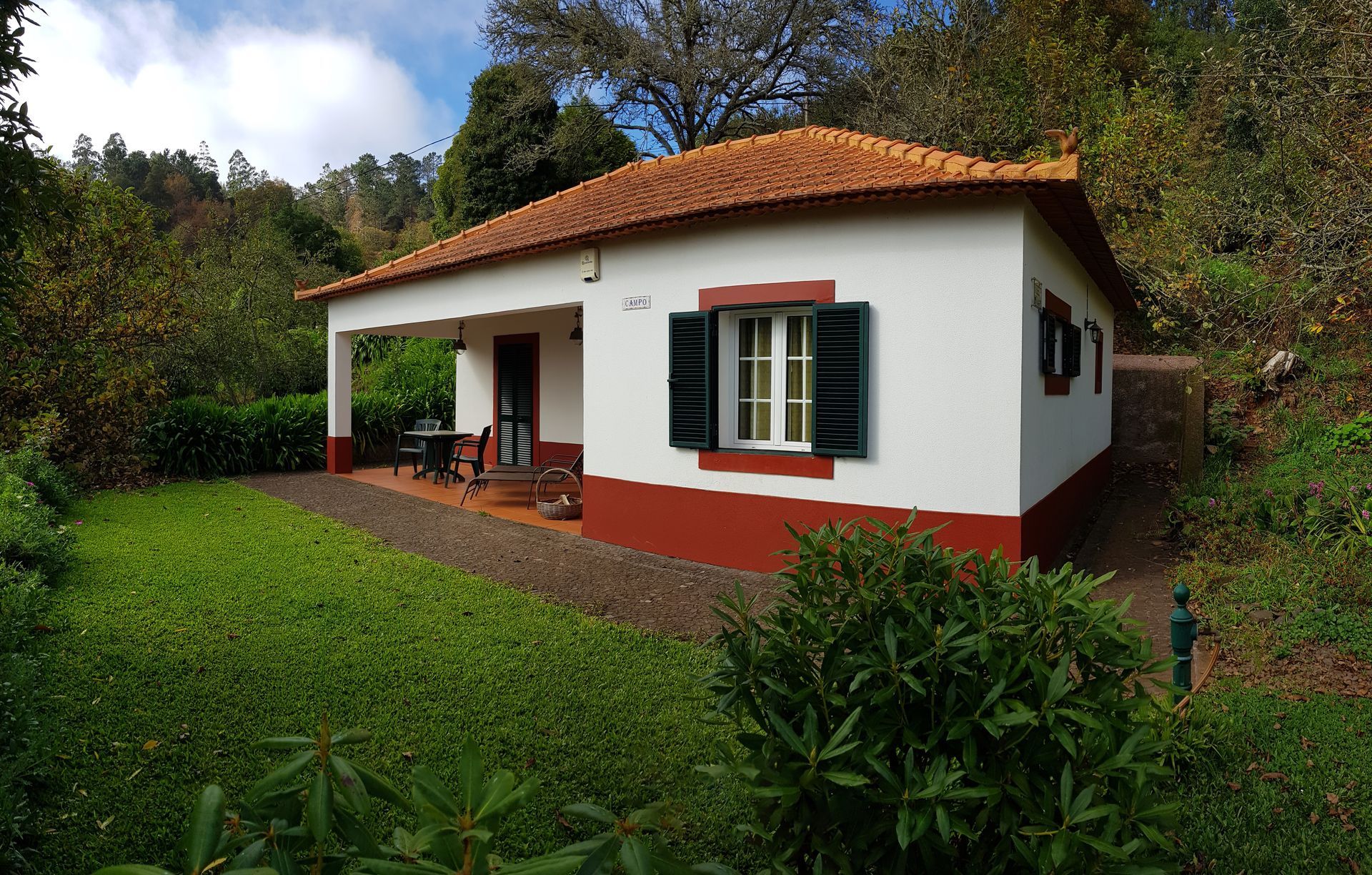 White cottage with red trim, terracotta roof, green shutters, and a small porch, surrounded by greenery.
