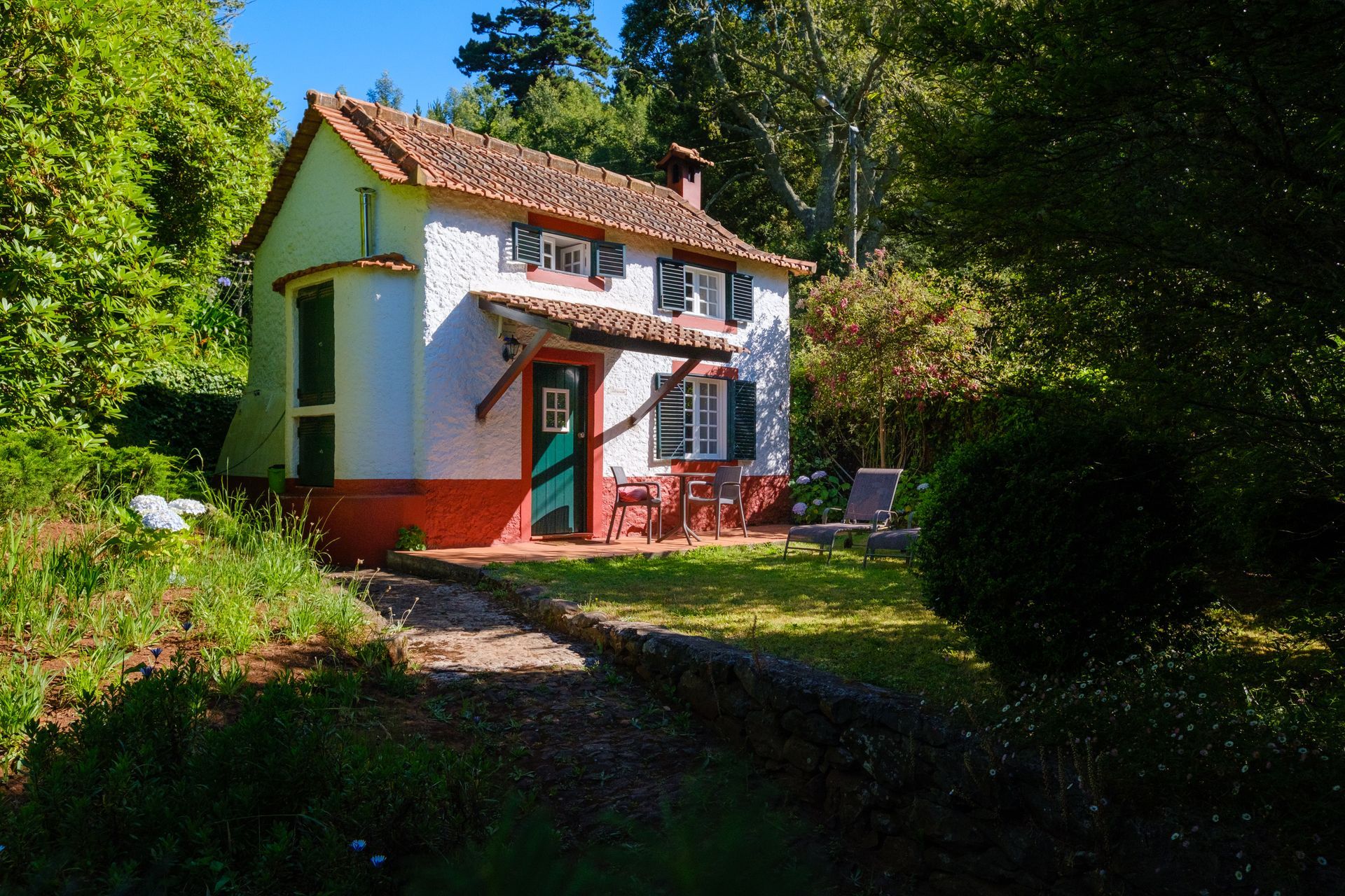 Quaint white cottage with red trim, surrounded by lush greenery, with a small lawn and patio.