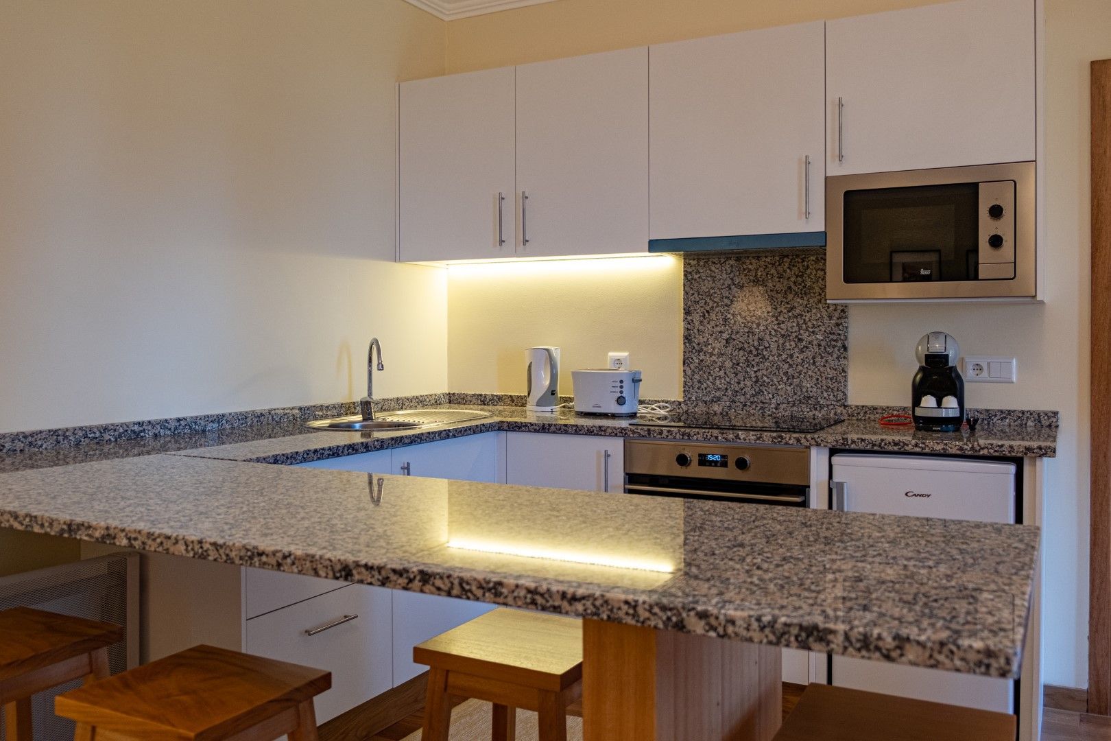 A modern, white kitchen with granite countertops, a microwave, and breakfast bar with stools.