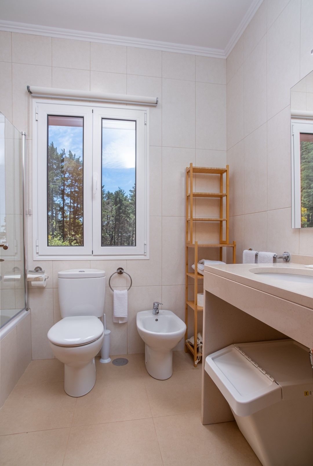 Bathroom with toilet, bidet, sink, and window overlooking trees. Light beige walls and floor.