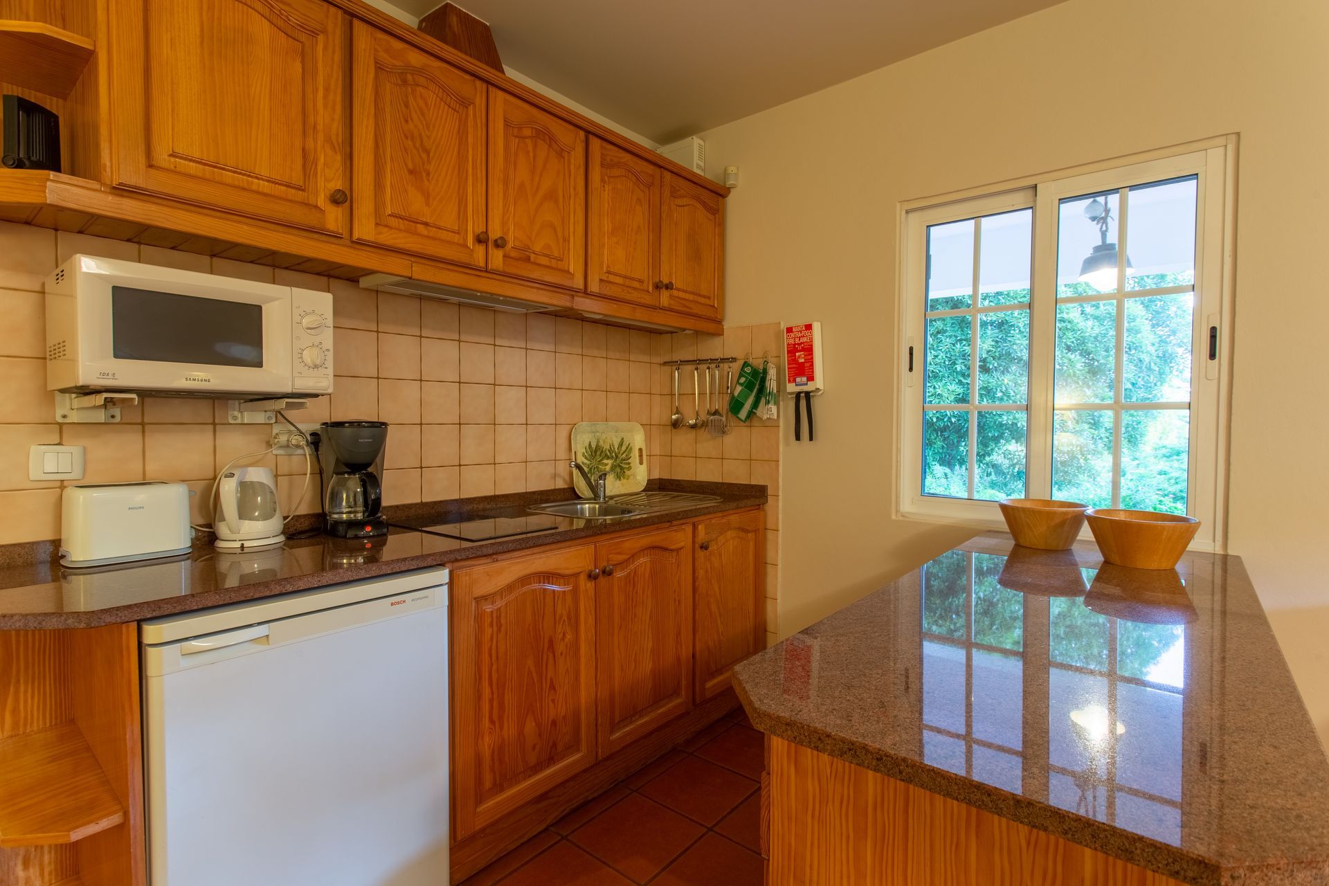 Kitchen with wood cabinets, microwave, refrigerator, and granite countertops. A window offers a view of greenery.