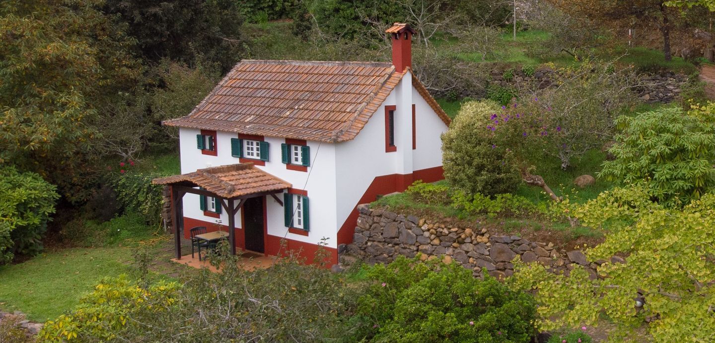 Small white house with red trim, surrounded by greenery, in a natural setting.