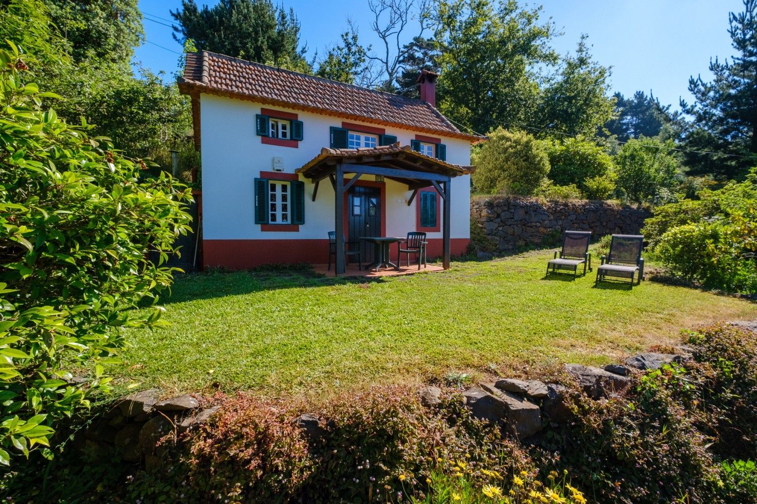 Small white cottage with red trim, green shutters, and porch in a lush green yard with lounge chairs.