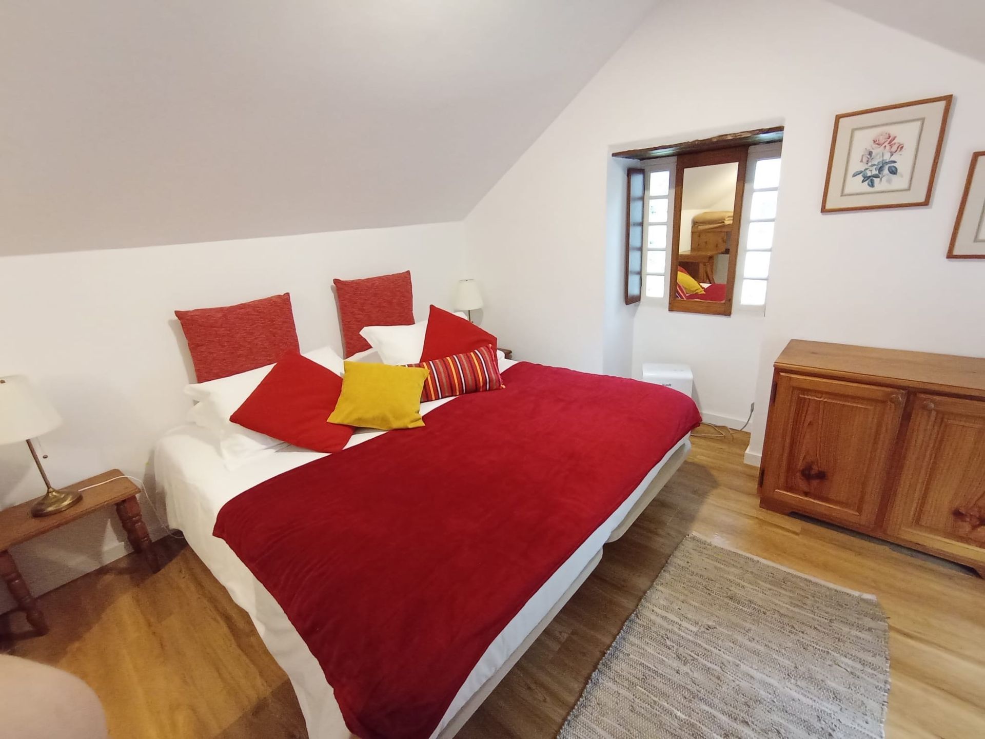 Bedroom with red bedspread, white walls, wooden furniture, and a small window.
