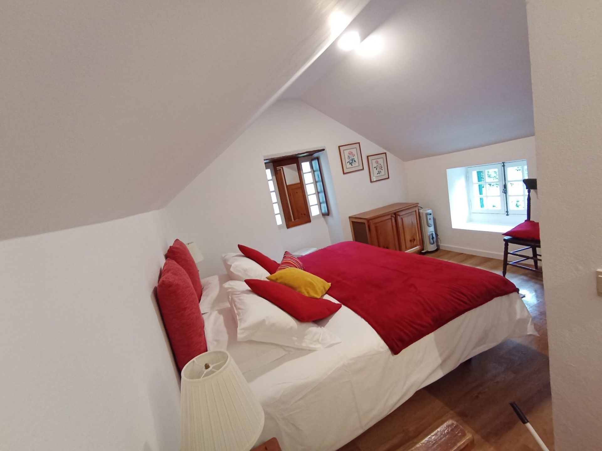 Bedroom with slanted white ceiling, bed with red bedding, small window, and wooden furniture.