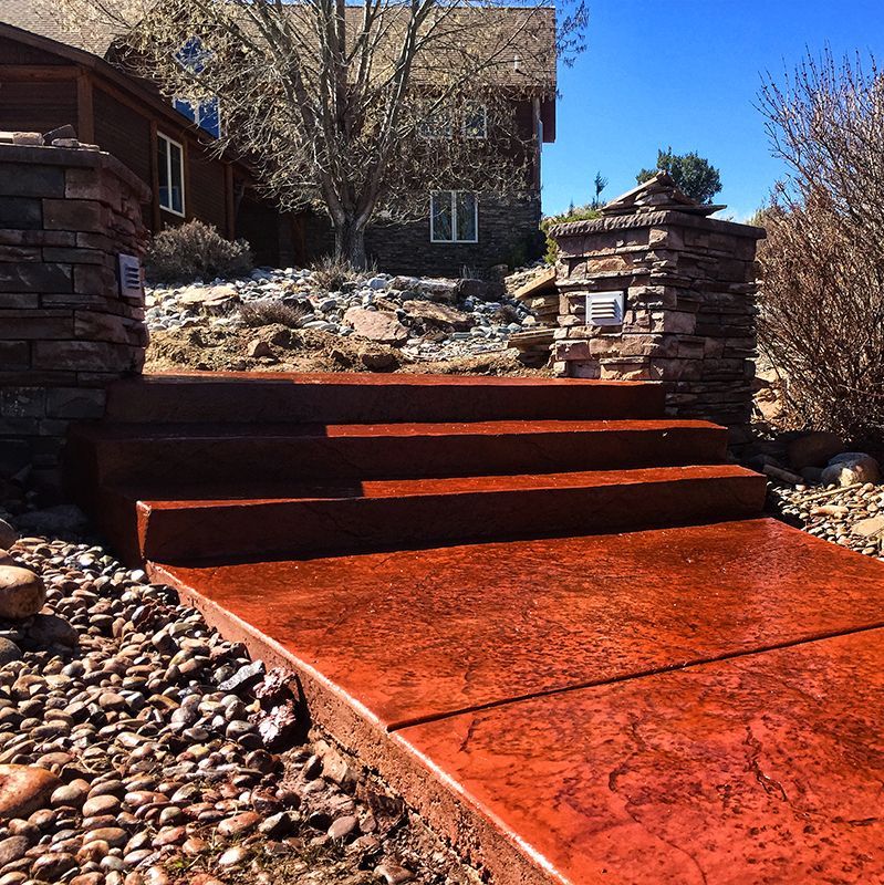 Red concrete walkway with steps leading up a landscaped front entrance between stone pillars.