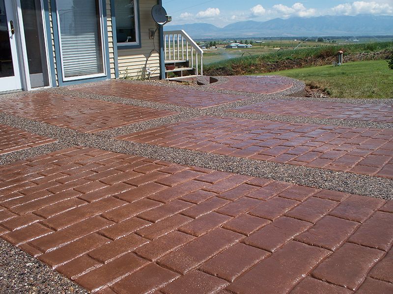 Red brick patio beside a house, overlooking a grassy landscape and distant water under a blue sky.