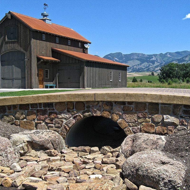 Stone bridge over a rocky stream with a rustic barn and mountain landscape under a clear blue sky