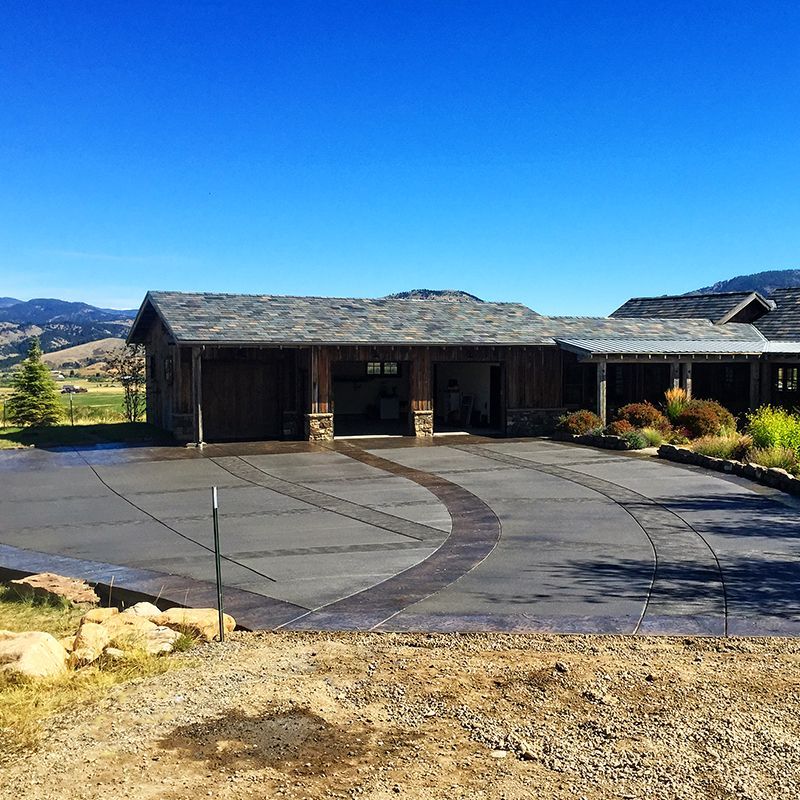 Modern rustic house with a circular driveway, stone exterior, and mountain landscape under a clear blue sky