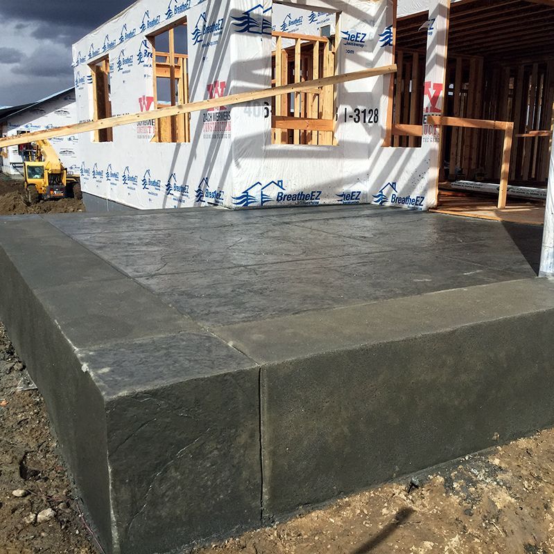 Freshly poured concrete slab foundation beside a partially built house with wood framing and windows.