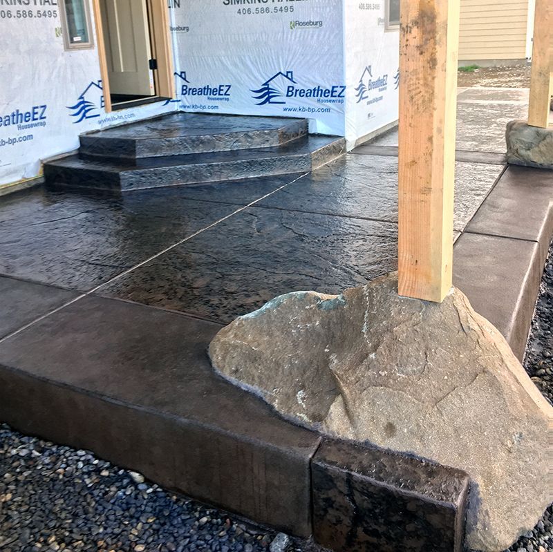 Concrete porch under construction with stone steps, wooden post, and house wrap visible