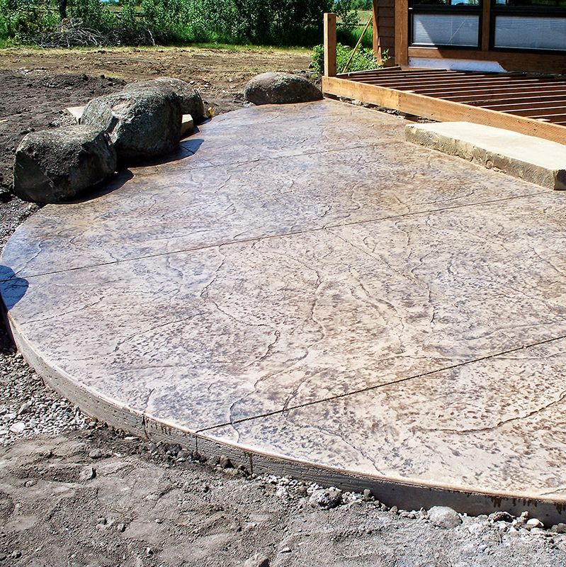 Curved stamped concrete patio beside a wooden deck, with large rocks and a house in the background