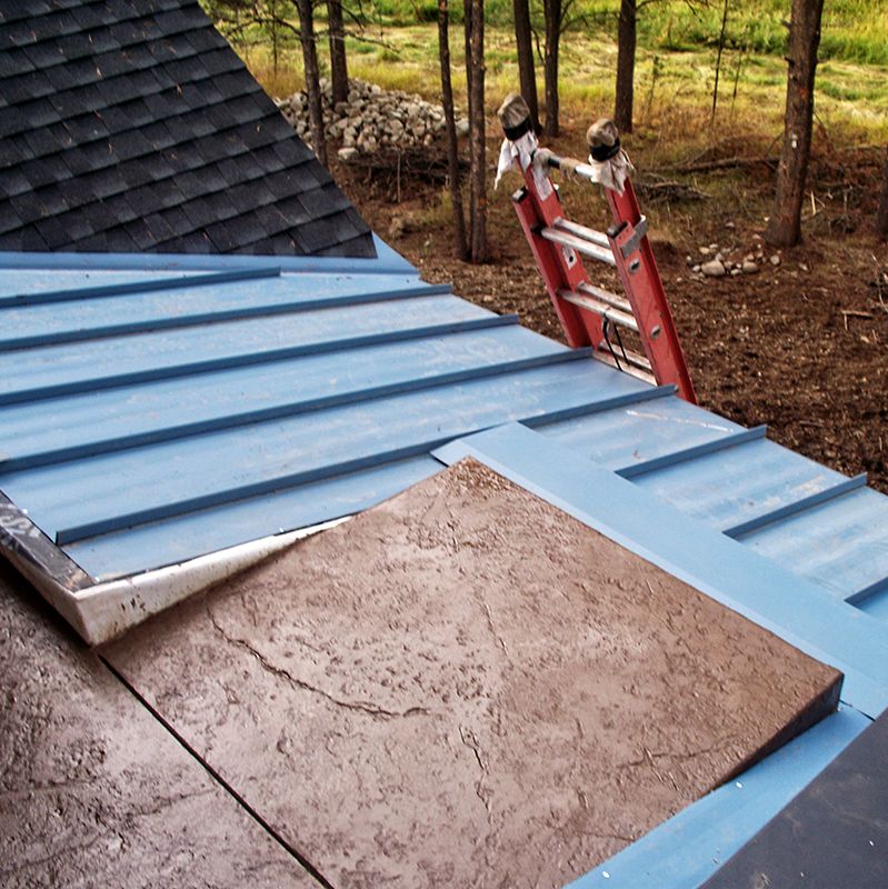 Blue metal roof with a concrete slab, viewed from above beside a ladder and wooded yard