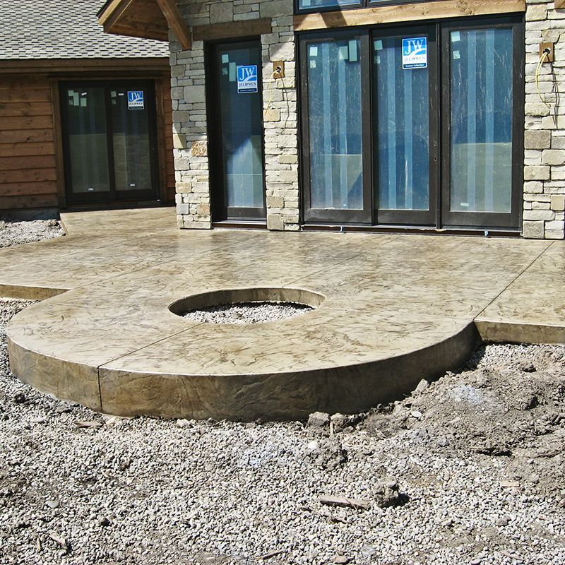 Concrete patio with circular fire pit in front of a house with large windows and stone siding