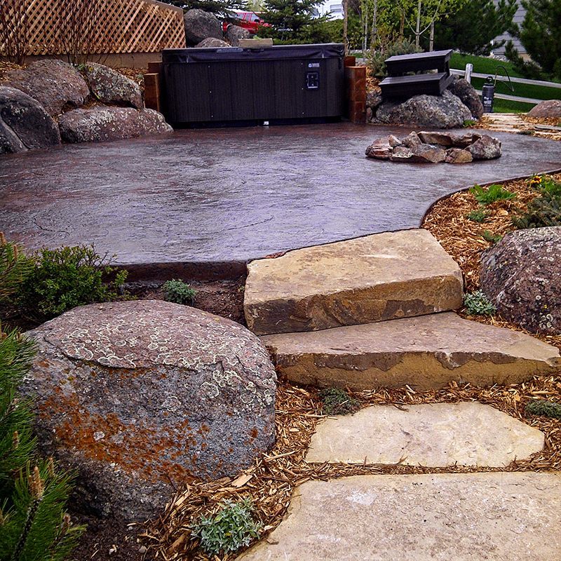 Curved stone steps leading to a wet paved patio with rocks and a black hot tub in a landscaped yard