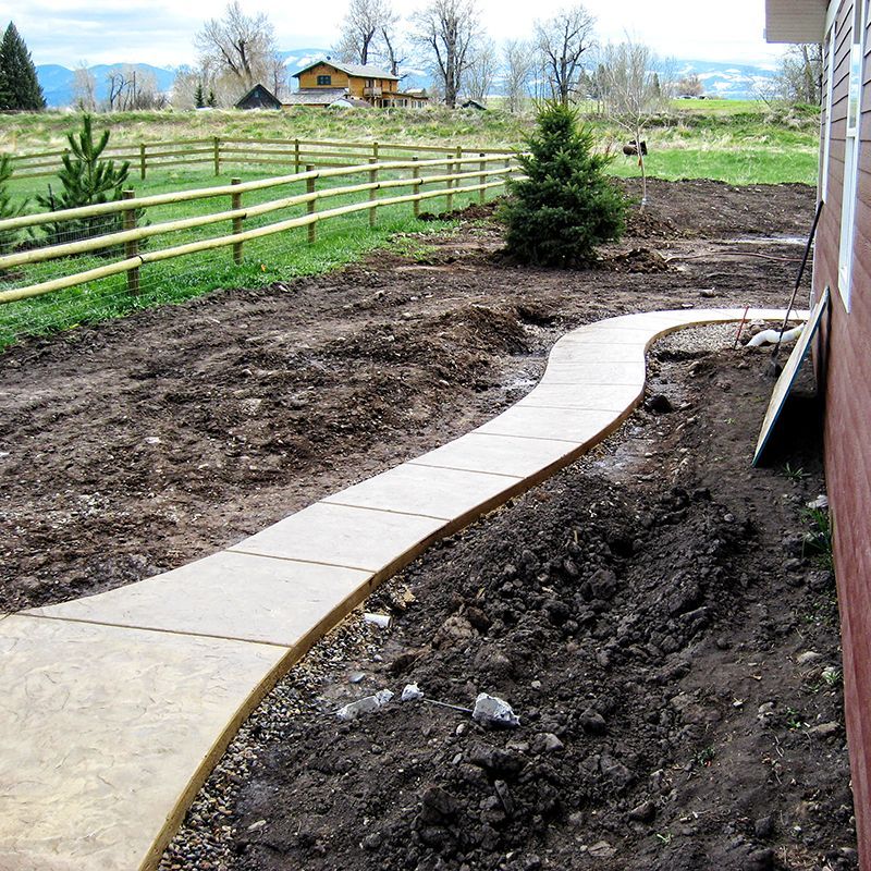Curved concrete garden path beside a house, bordered by fresh soil and a wooden fence in a rural yard.