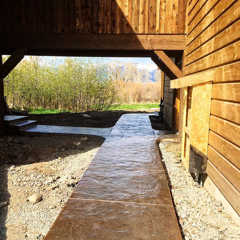 Covered stone walkway beneath a wooden overhang beside a cabin, leading to a sunny yard and trees.