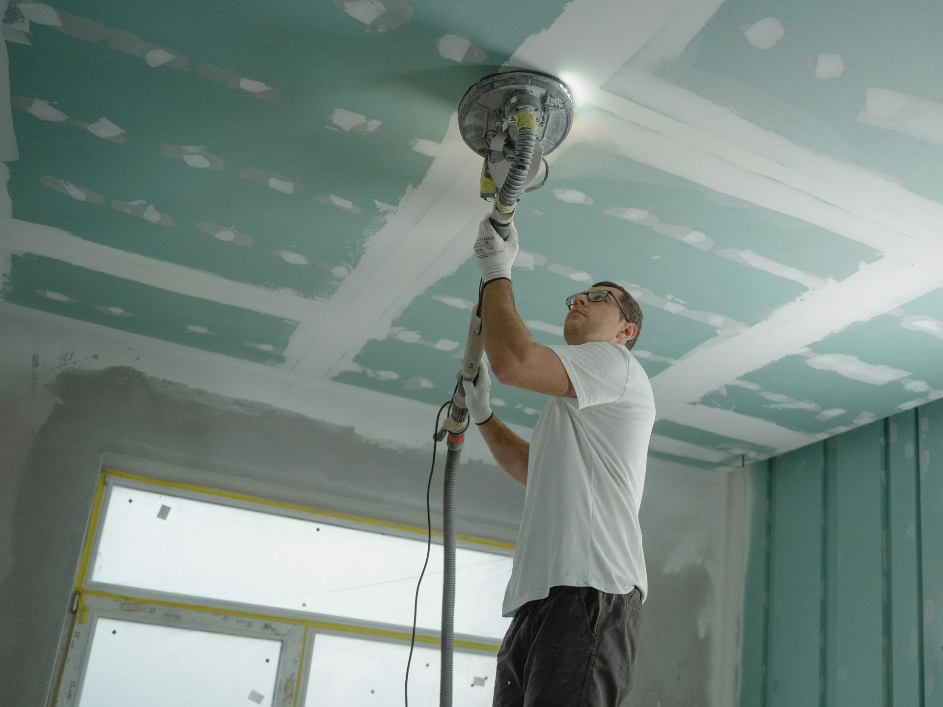 Man sanding a drywall ceiling with a long-handled sander, in a room with green drywall.