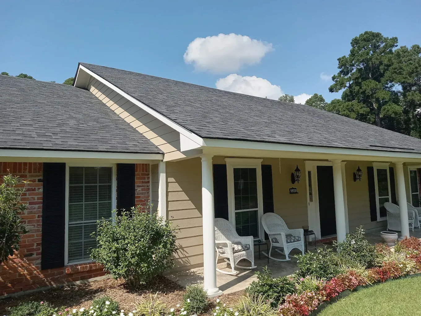 A house with a gray roof, brick facade, and beige siding, with white rocking chairs on the porch.