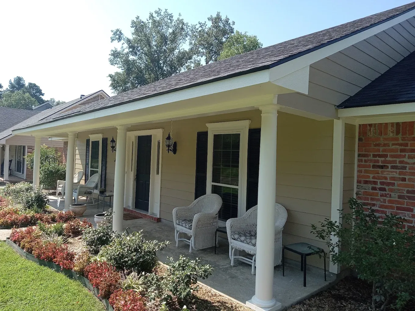 Exterior of a beige house with a porch, white columns, and a red brick accent. Flowers and chairs visible.