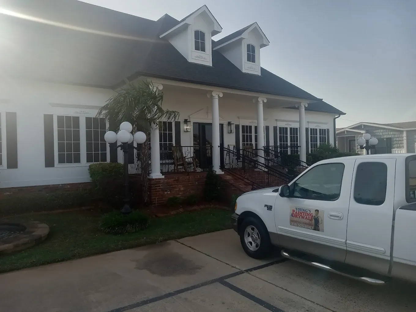 White house with black shutters and a white truck in the driveway on a sunny day.