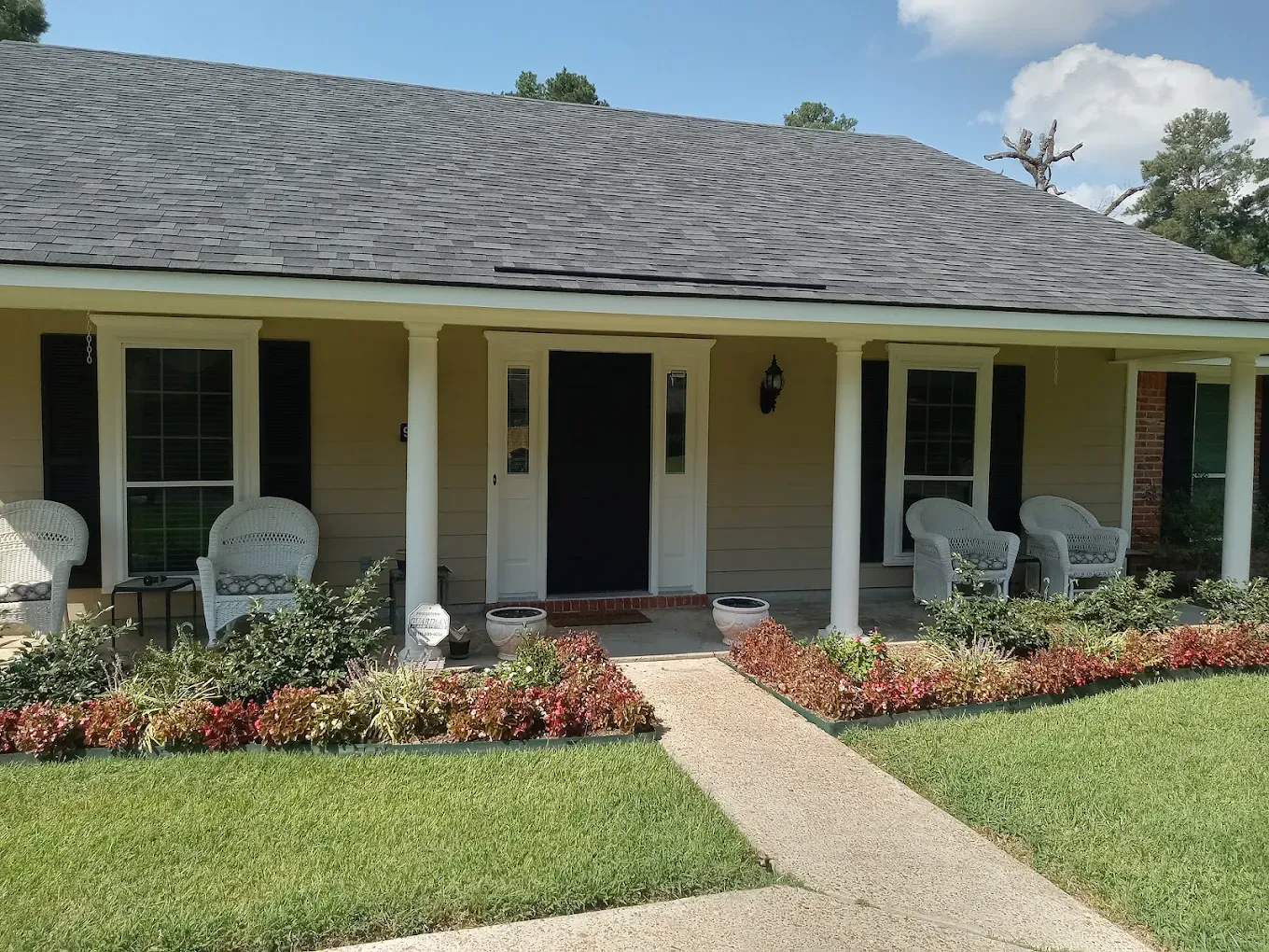 Tan house with porch, white pillars, shutters, wicker chairs, red and green landscaping, and a concrete walkway.