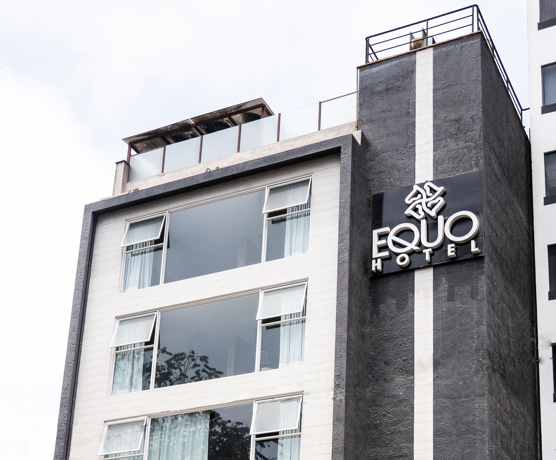 Edificio gris y blanco de varios pisos con reloj, autos estacionados en el frente, nublado.
