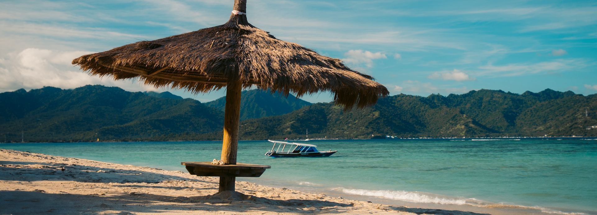 Beach scene with a thatched umbrella, turquoise water, and green mountains in the background.