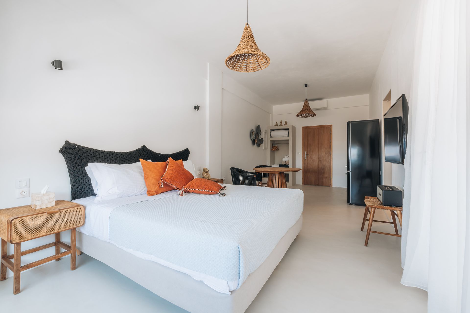 Bright bedroom with a bed, nightstand, TV, and a dining area. Mostly white with wooden accents and orange pillows.