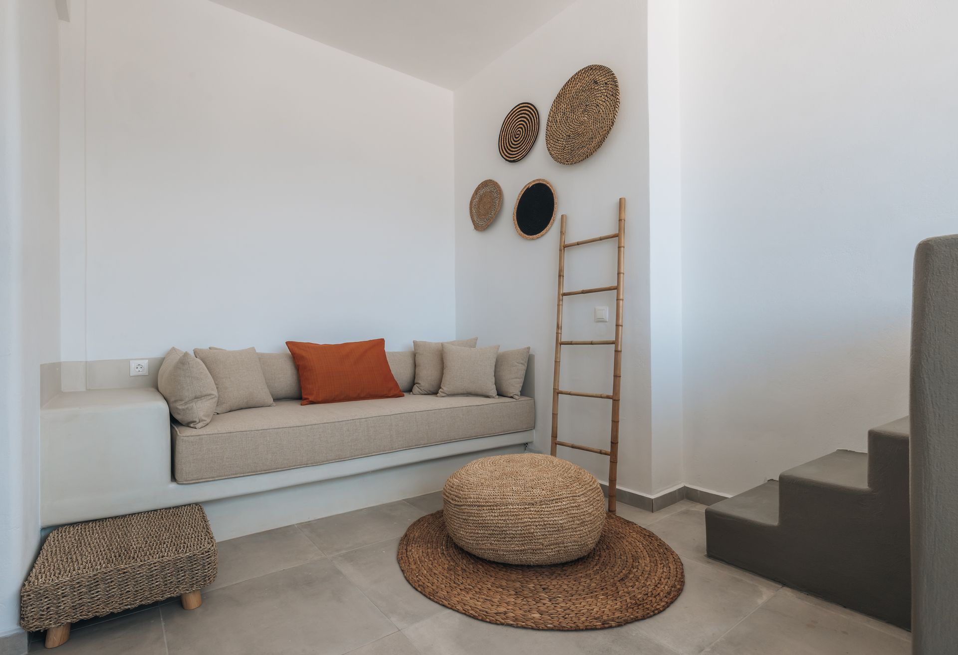 Cozy, white-walled seating area with built-in couch, woven decor, small stool, pouf, and ladder.