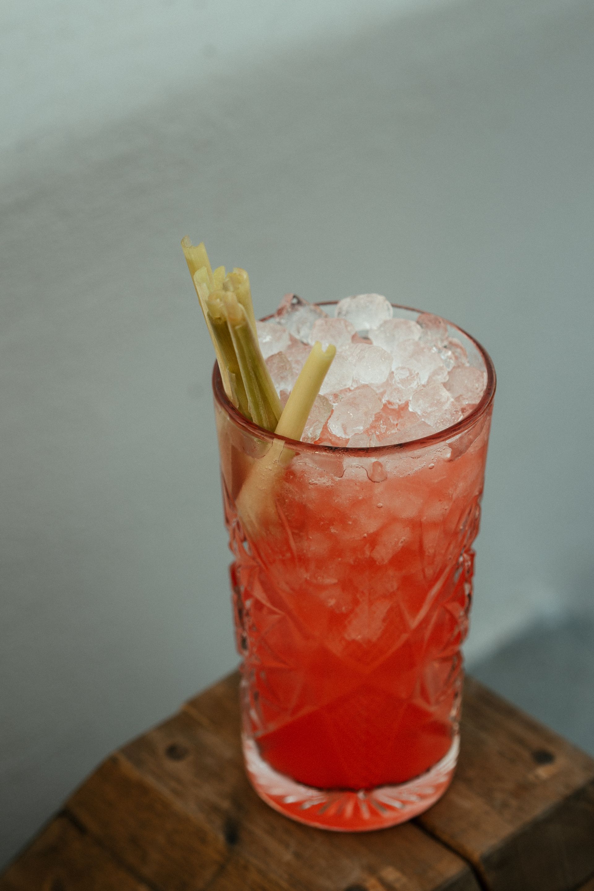 Red cocktail in a patterned glass with crushed ice and lemongrass stalks on a wooden surface.