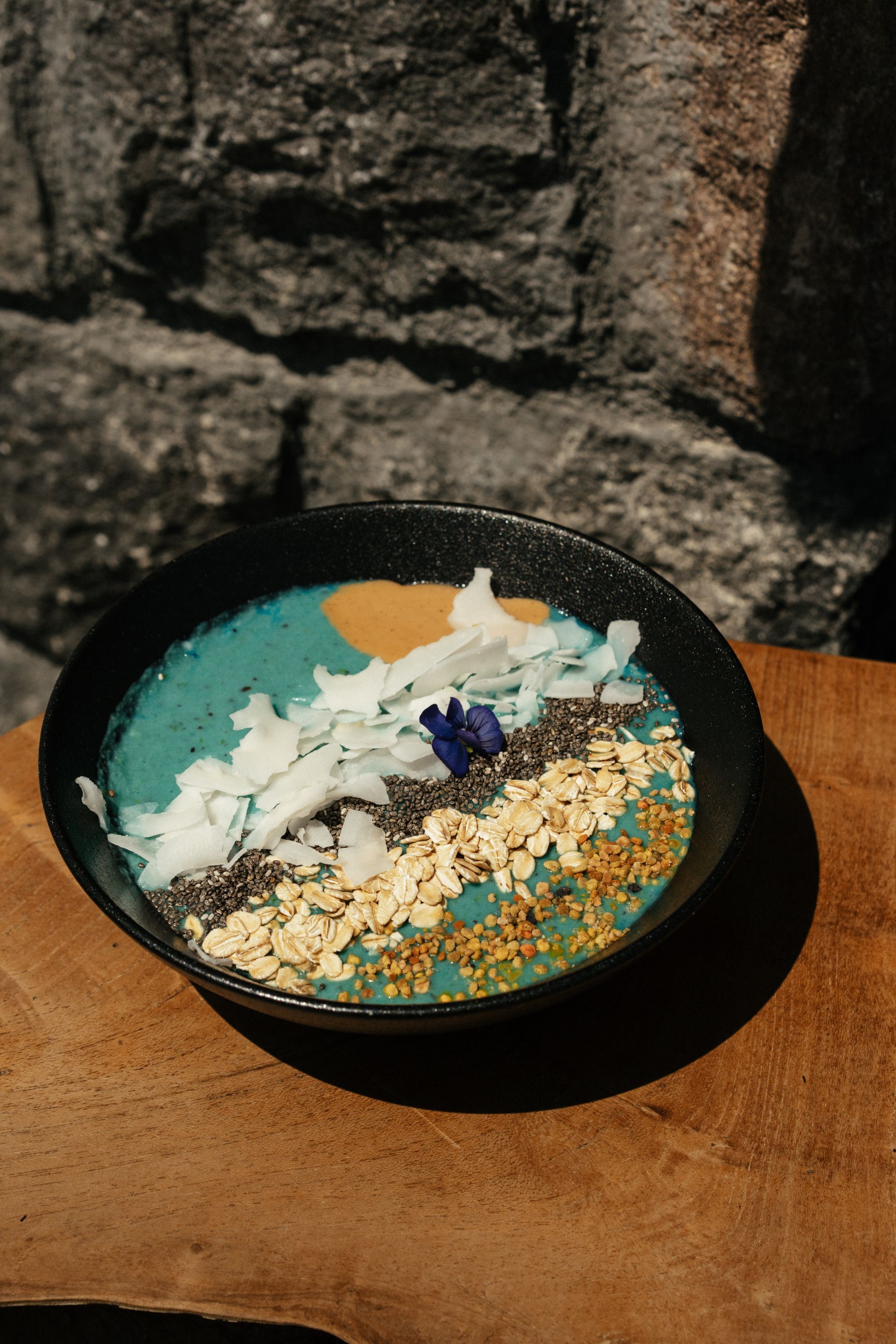 Blue smoothie bowl with toppings on a wooden surface, against a stone wall.