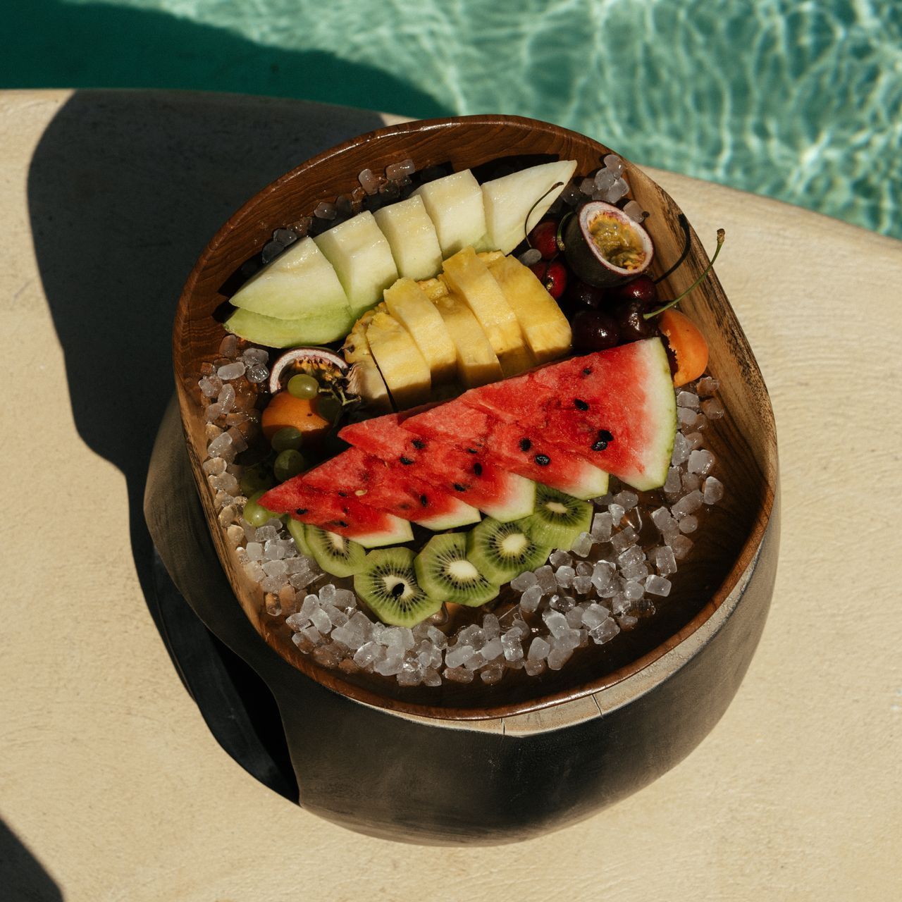 Fruit platter with watermelon, kiwi, pineapple, and melon on ice near a pool.