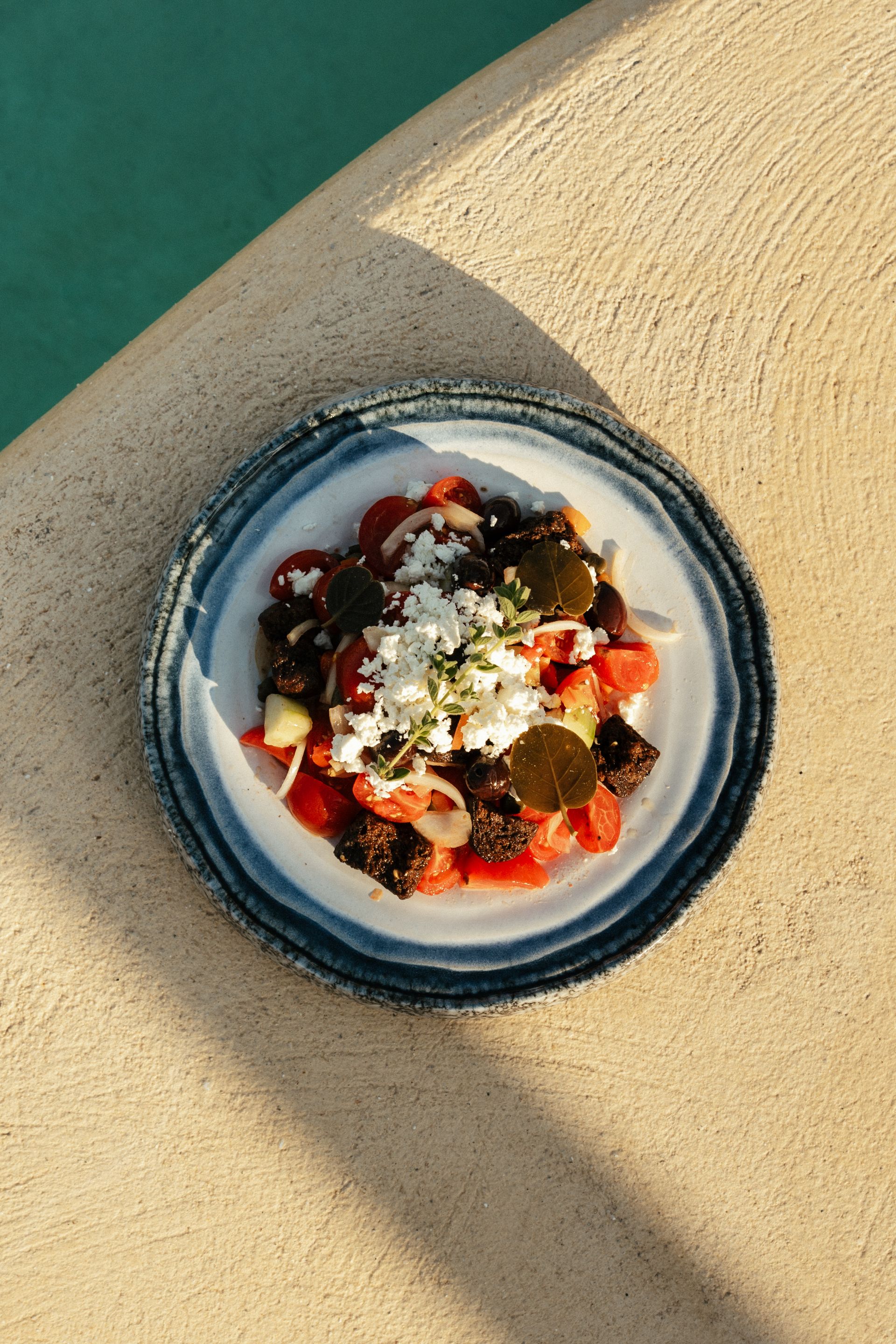 Salad with tomatoes, olives, and feta cheese on a blue-rimmed plate, near a pool.