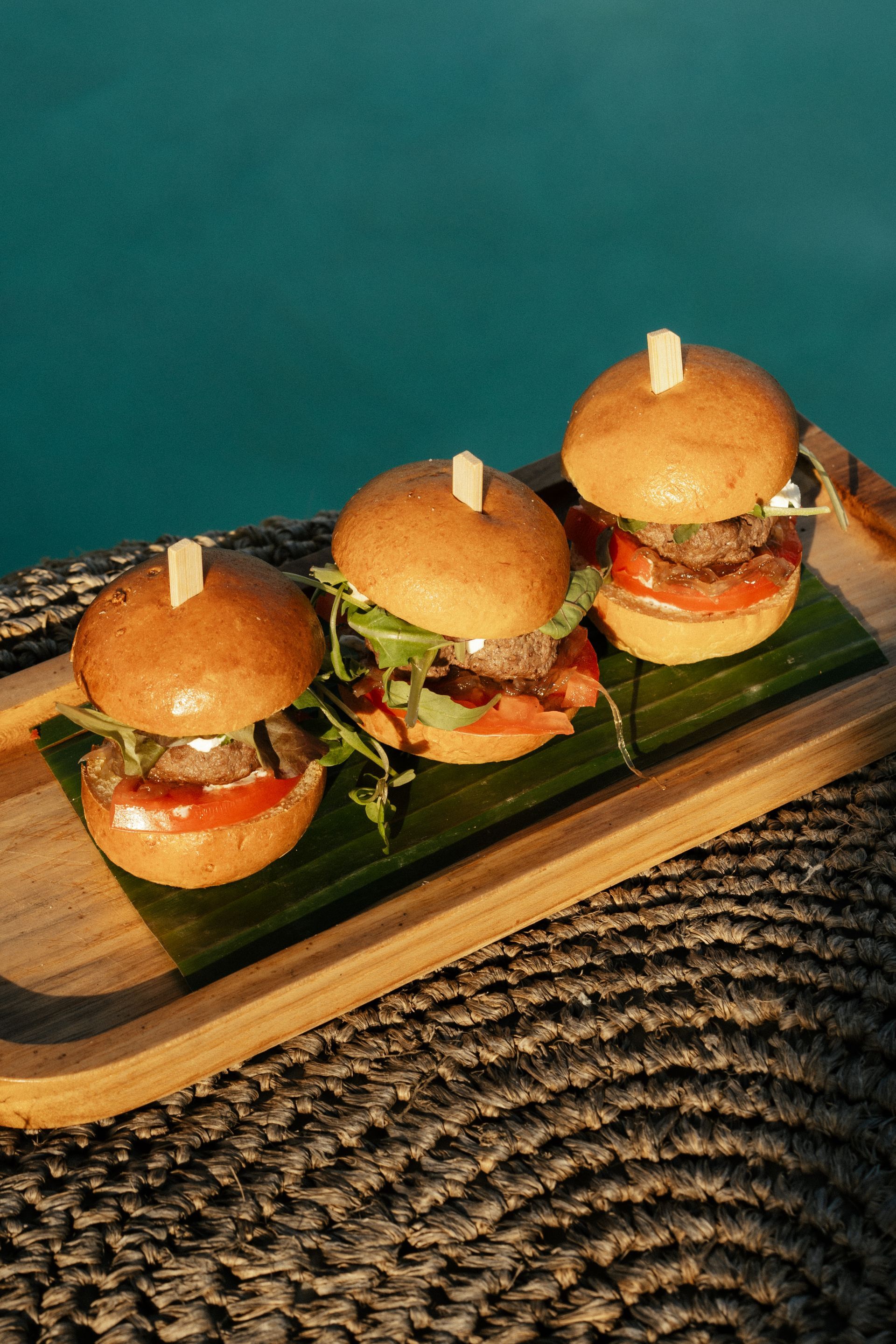 Three mini burgers on a wooden tray, resting on a banana leaf, near a pool.