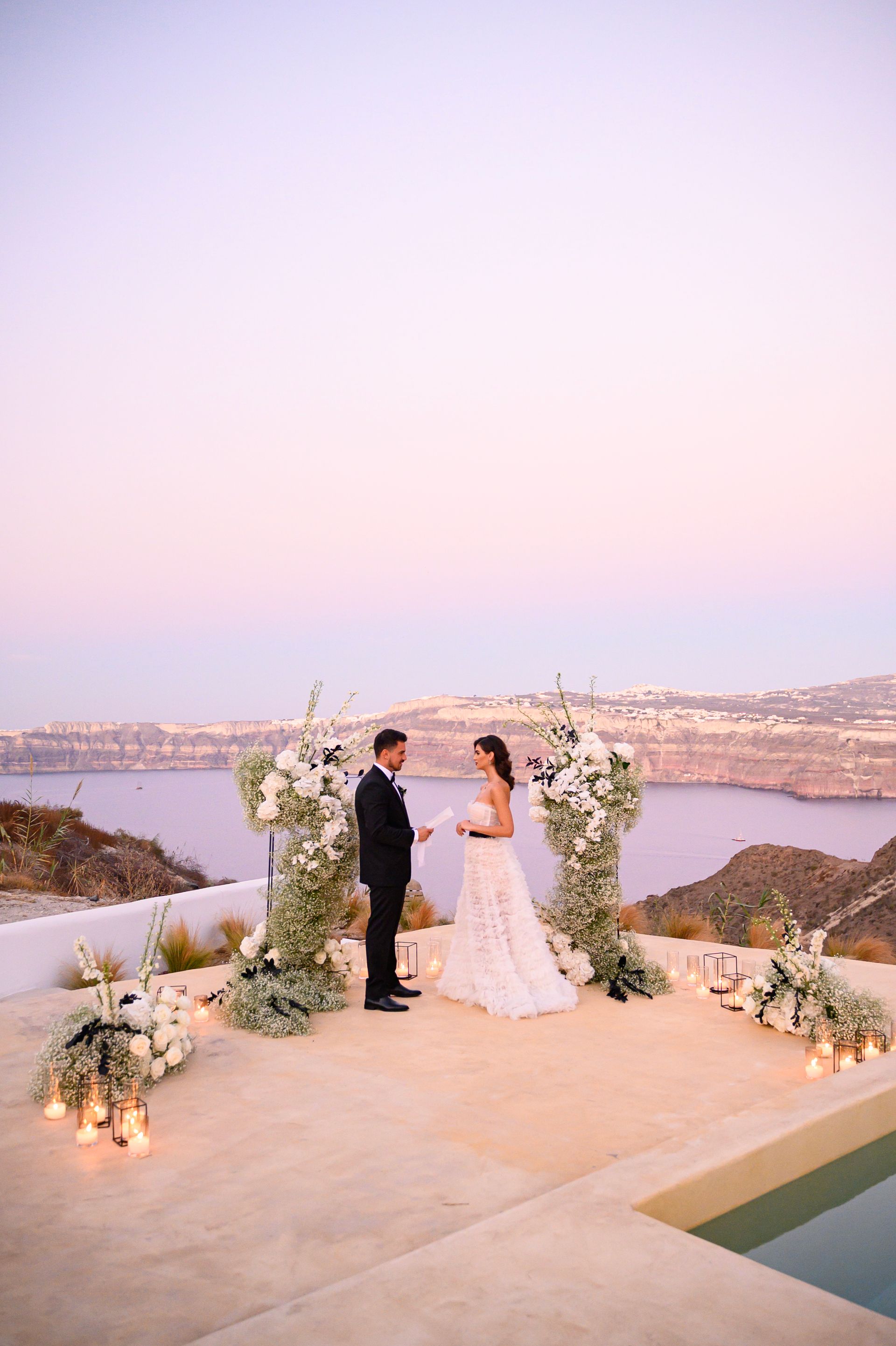 Couple exchanges vows under floral arch overlooking ocean at sunset.