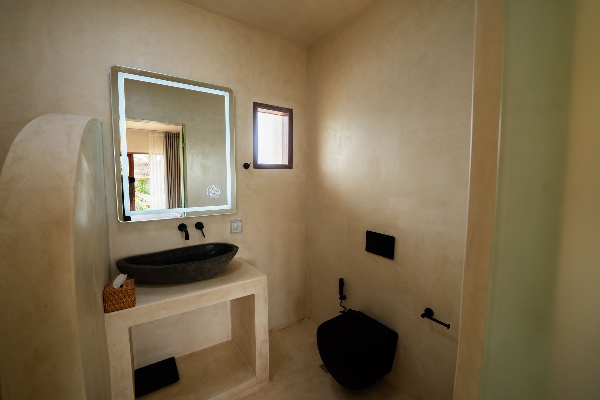 Bathroom with black toilet and sink, illuminated mirror, cream walls, and small window.