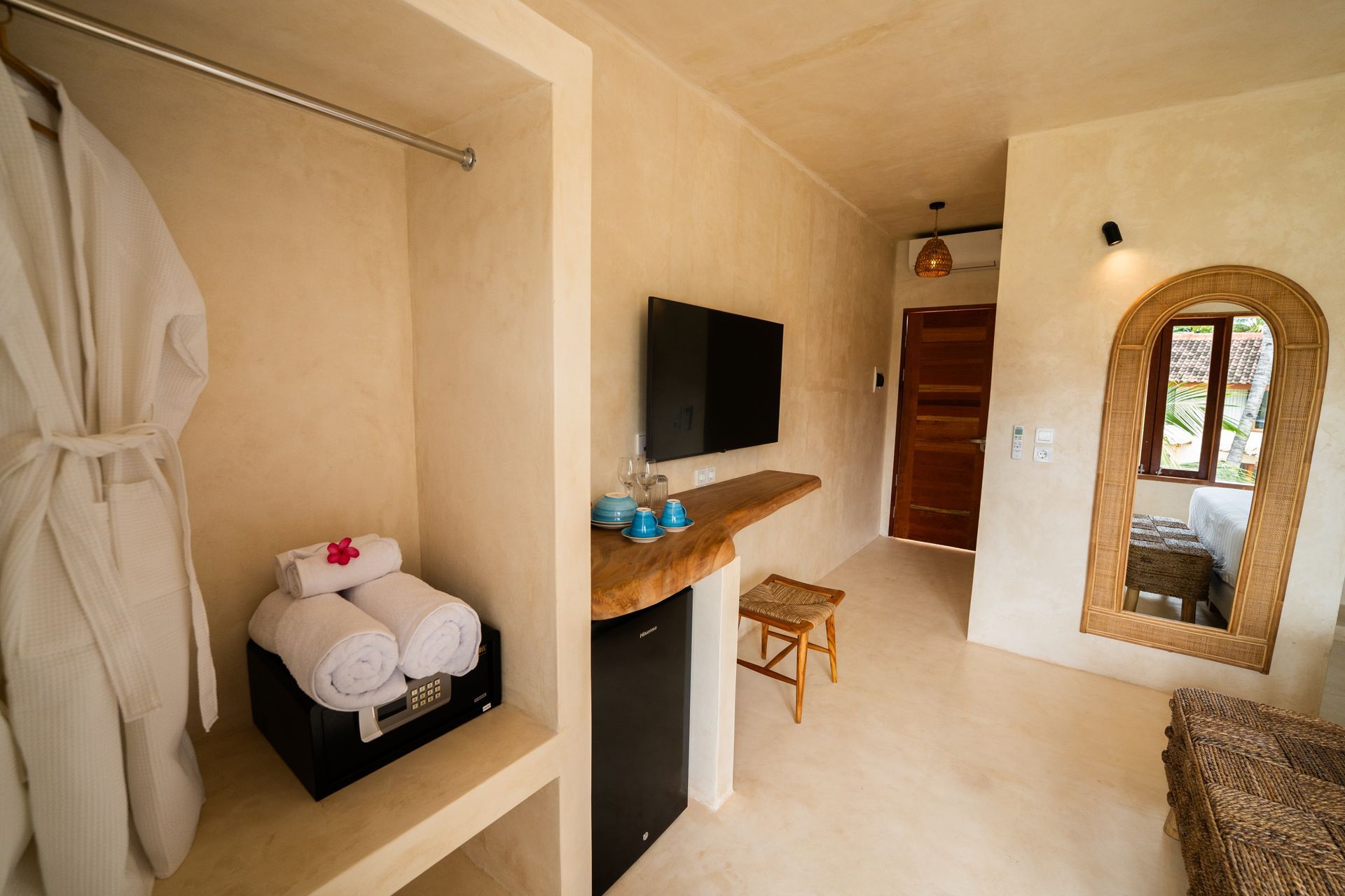 Hotel room interior: wardrobe with robe, towels, TV, mirror, stool, light-colored walls, and floor.