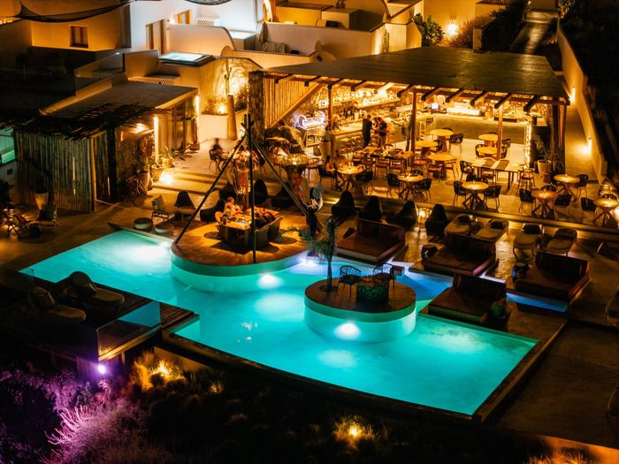 Nighttime view of a pool area with illuminated water and a restaurant with people; white buildings in the background.