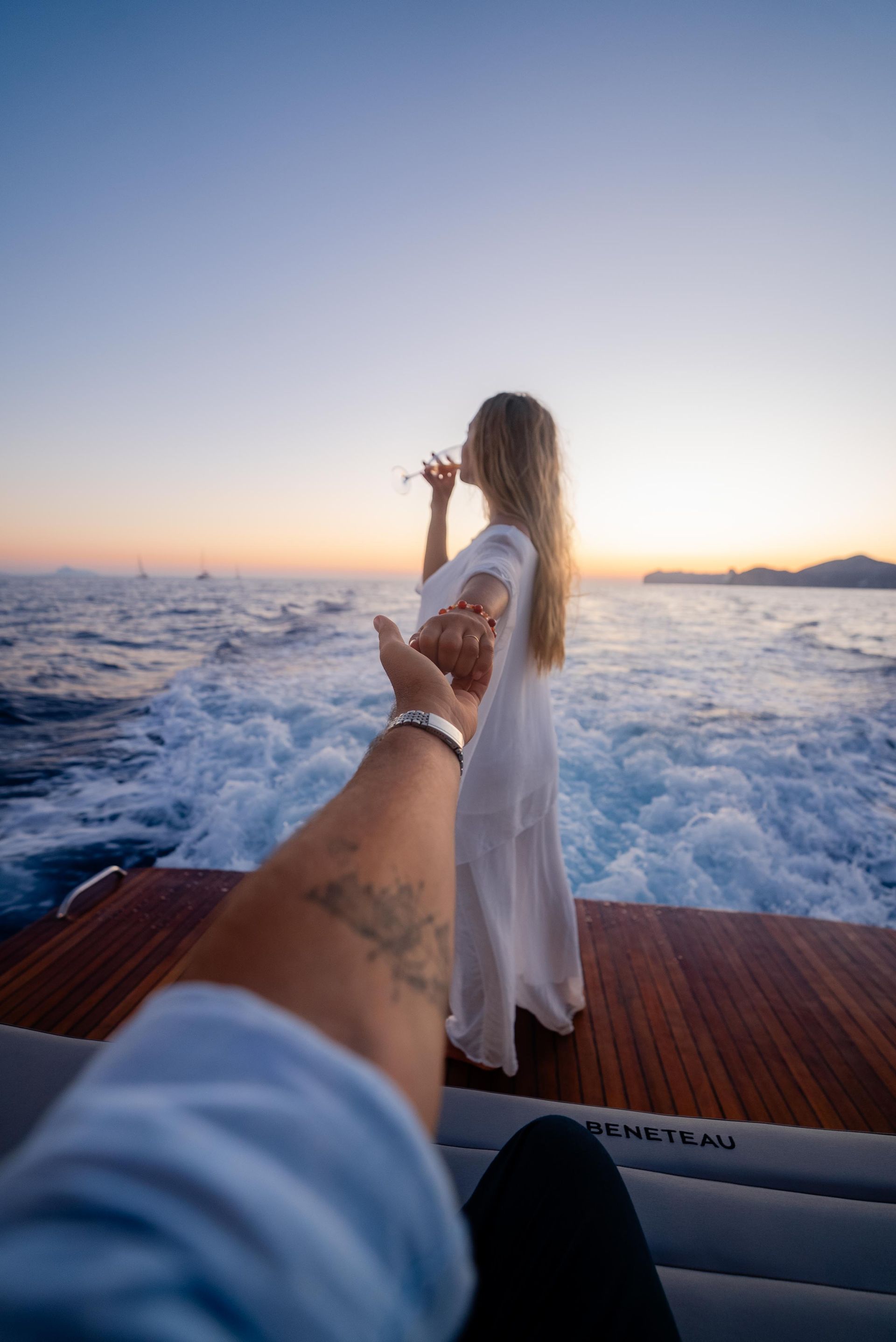 A person holding hands with a woman on a boat, drinking wine, at sunset over the ocean.