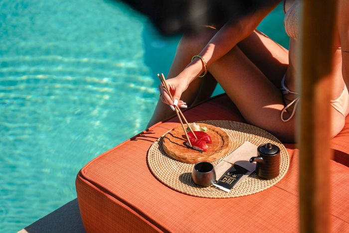 Woman eating cake by a pool. Wearing a bikini, she uses chopsticks. The setting is outdoors, sunny, and relaxing.