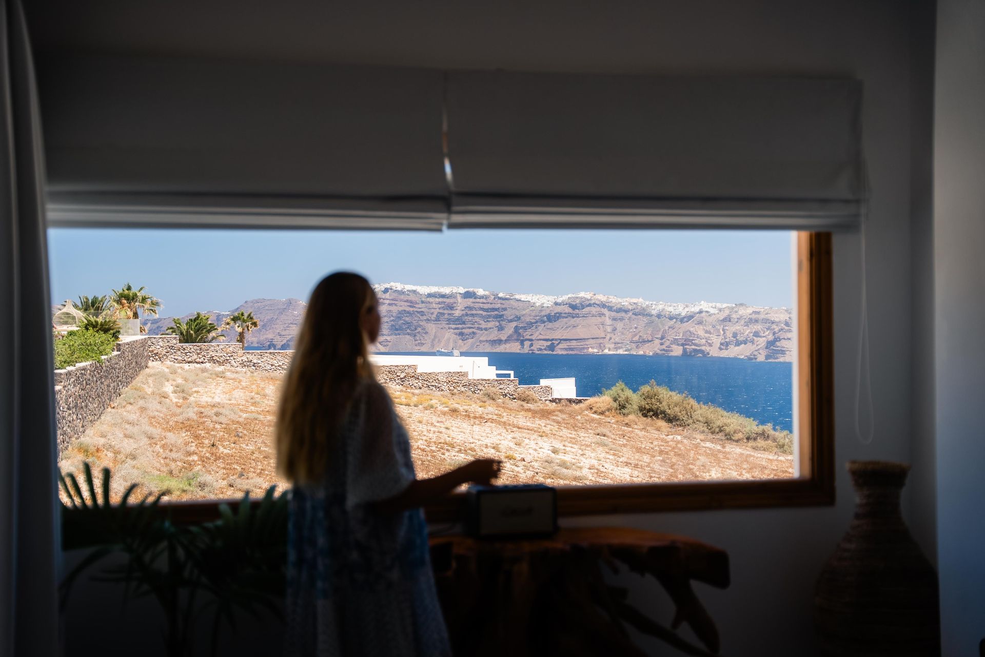 Woman looking out a window at a coastal landscape with white buildings and blue water on a sunny day.