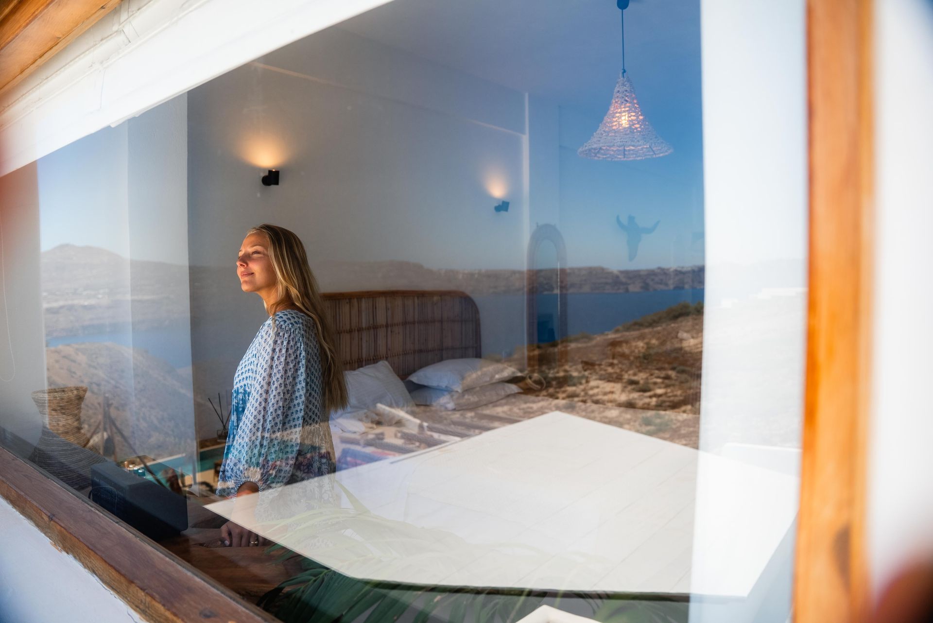 Woman looking out a window at the sea from a white room with a bed.