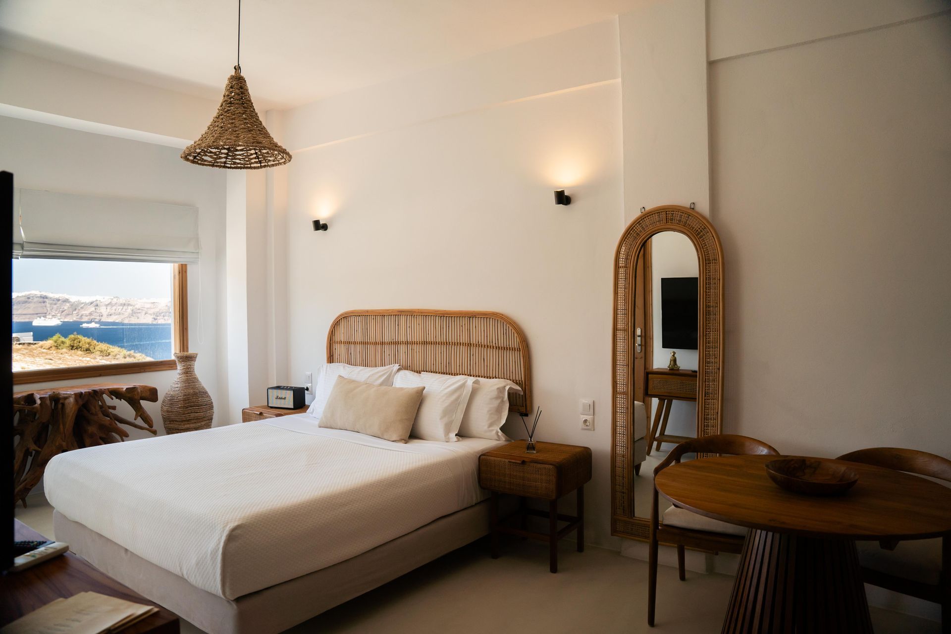 A bright white bedroom with a bed, wicker accents, round table, and a view of the sea.