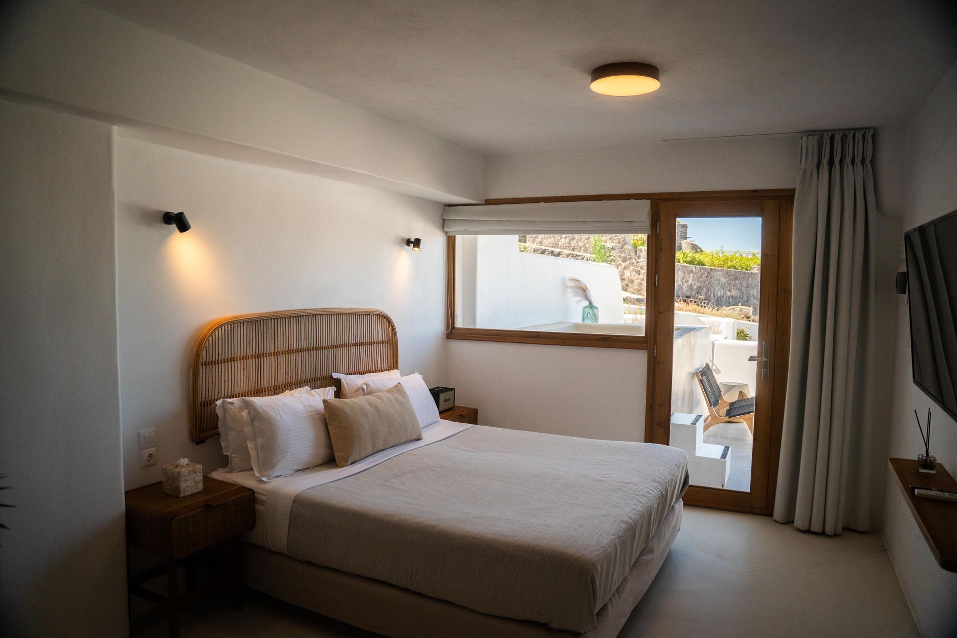 Bedroom with a bed, window, door, and a rattan headboard. Off-white walls and bedding, wooden accents.
