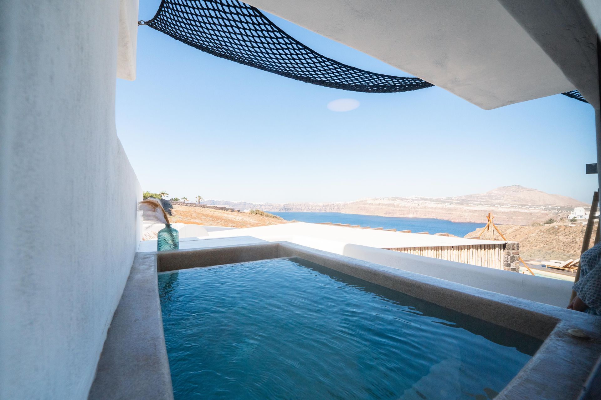 Outdoor plunge pool overlooking a blue sea, shaded by a net, with white walls.
