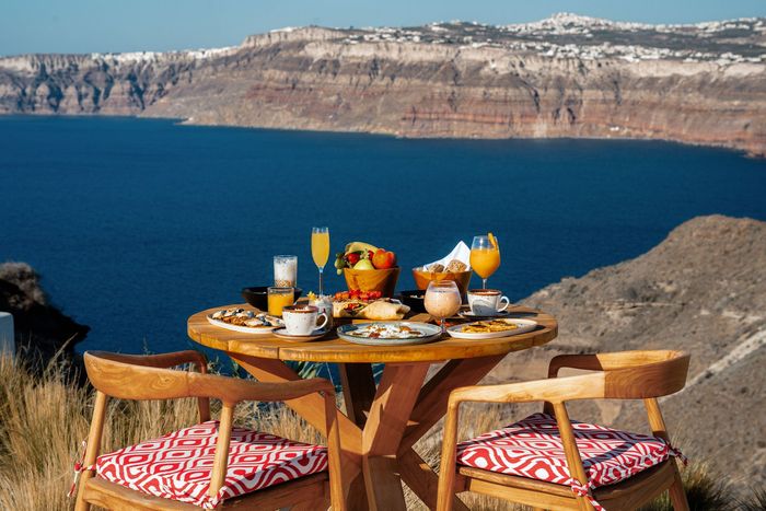 Breakfast table overlooking blue sea and cliffs, with food, drinks, and chairs.