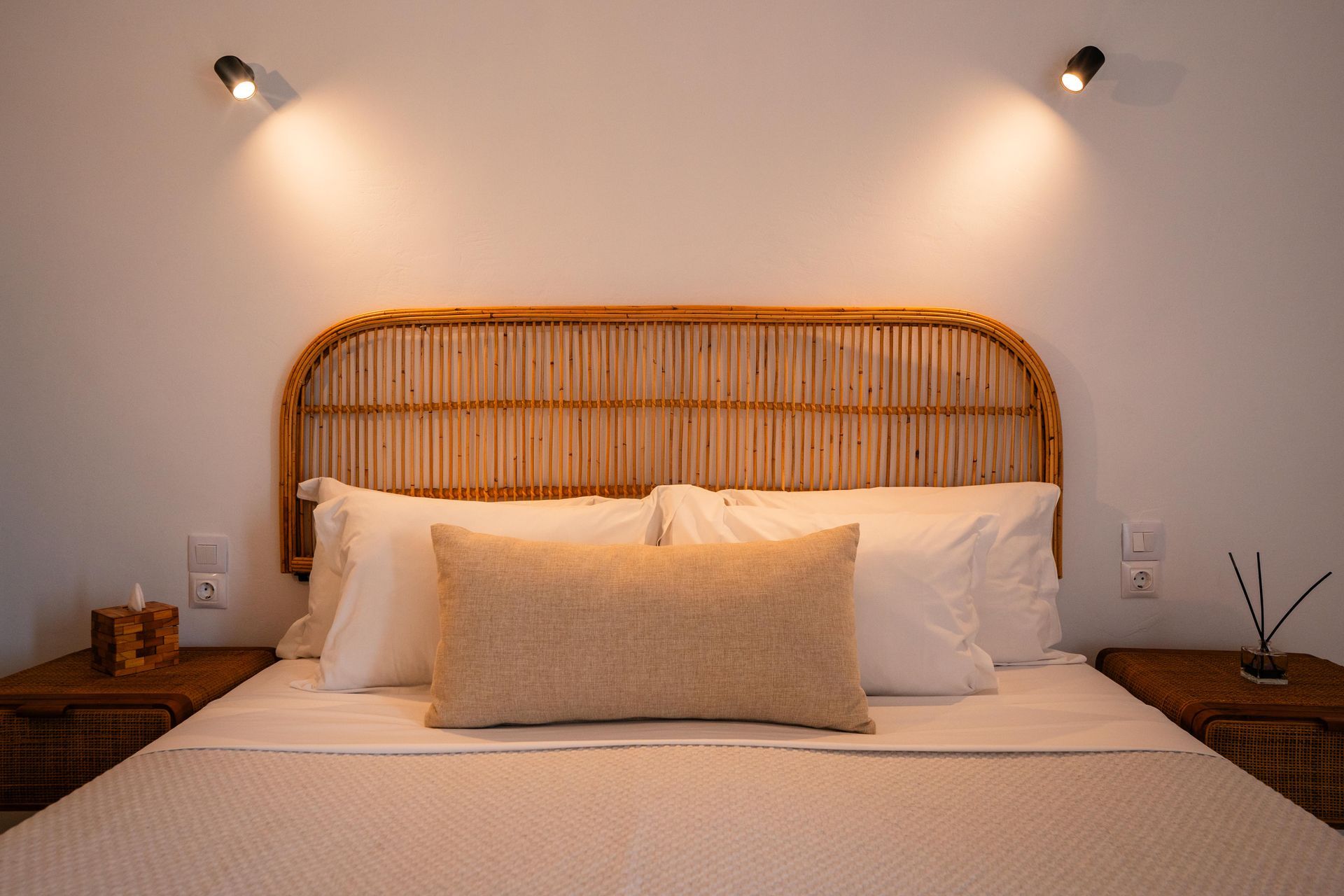 Bedroom with rattan headboard, two bedside tables, and two spotlights illuminating the bed.