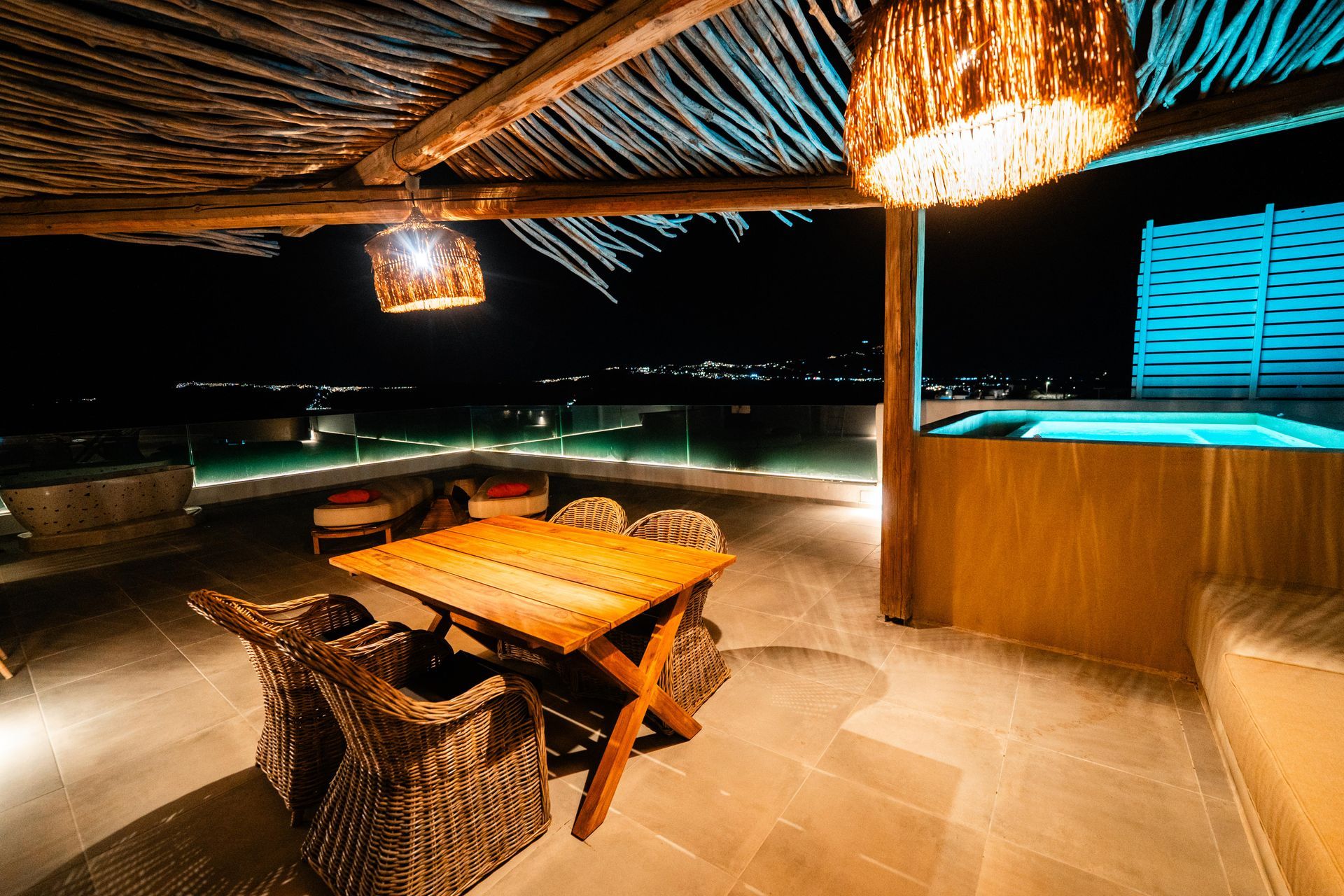 Outdoor patio at night with a wooden table and chairs, lit by hanging lamps, with a hot tub and a view of the ocean.